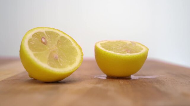 Slow motion horizontal video of two lemon halves resting on a wooden board as a hand enters at the end of the shot and removes one half against a white background