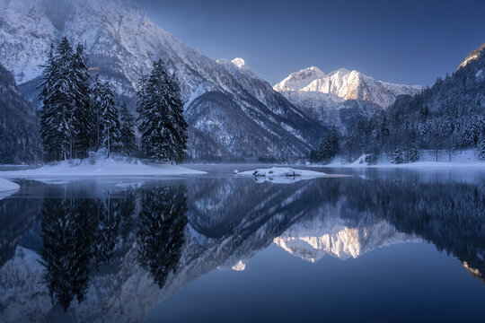 View of snow-covered mountains pierce the crisp blue sky, mirrored in the serene lake below, creating a tranquil winter scene, Tarvisio, Friuli-Venezia Giulia, Italy.