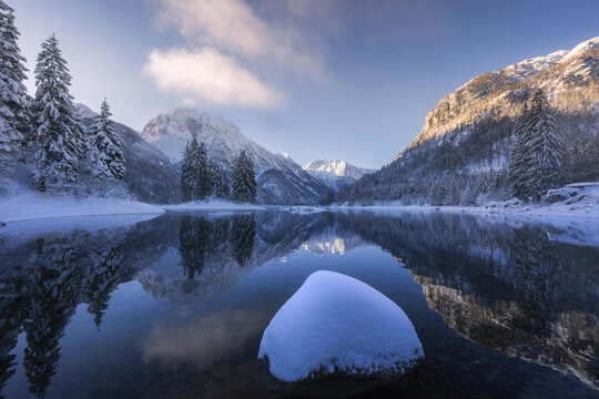 View of snow-laden evergreens and majestic mountains mirror in the tranquil waters, a serene winter landscape, Tarvisio, Friuli-Venezia Giulia, Italy.