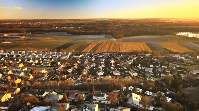 Drone descends smoothly over Elchingen at golden hour, lowering toward village rooftops with patchwork farmland and distant forest under warm sunset light.