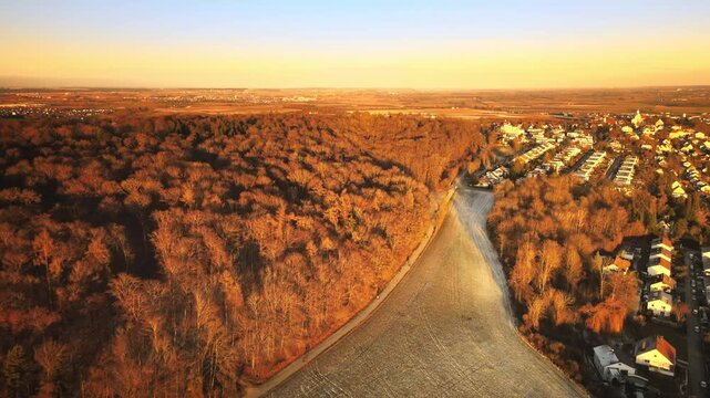 Forward-moving drone at golden hour near Elchingen, flying over a frosty field beside a dense forest and approaching residential rooftops, with warm light and long shadows.
