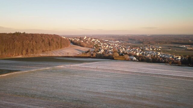 Forward-moving drone flight over frosty agricultural fields toward Elchingen, with forest edge on the left and a clear winter sky over the distant village and valley.