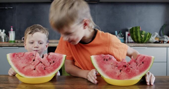 Playful boy leans forward with watermelon wedge as another boy sits near bowl. Child takes giant bite tugging rind hard while friend looks down keeping chewing