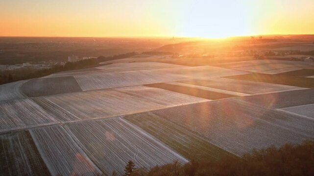 Forward-moving drone flight over frost-covered patchwork fields toward a bright sunrise, with cinematic lens flare, soft haze, and rolling farmland stretching to the horizon.