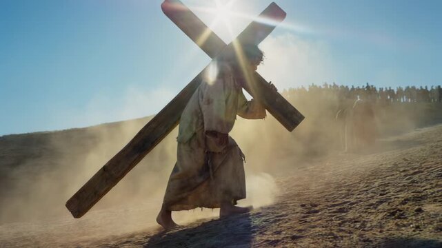 Jesus Christ carrying a heavy wooden cross up a dusty hill towards Golgotha. Biblical scene of the Passion and crucifixion with a crowd in background. Christian religious Easter concept