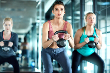 Fototapeta premium Women, fitness or weightlifting with kettlebell in gym for squat, workout or exercise together. Active, female people or group lifting with weight for muscle gain or strength training in health club