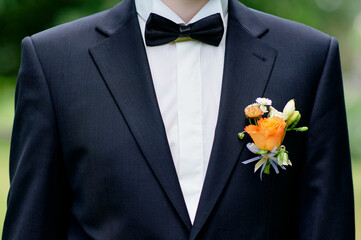Stylish groom wearing a classic black tuxedo with a vibrant orange boutonniere, captured outdoors against a soft green background. Perfect for wedding, celebration, or fashion concepts.