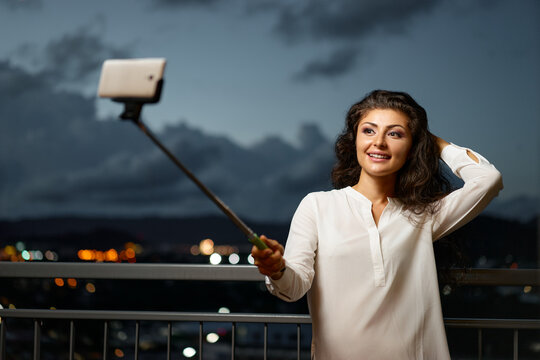 A cheerful woman in a white blouse poses for a selfie on a balcony at dusk, holding a selfie stick. City lights glow in the background, creating a warm, candid moment.