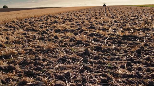 The field shows a wide expanse of tilled earth, with straw remnants scattered across the ground. A tractor plows the field, preparing the land for future crops.