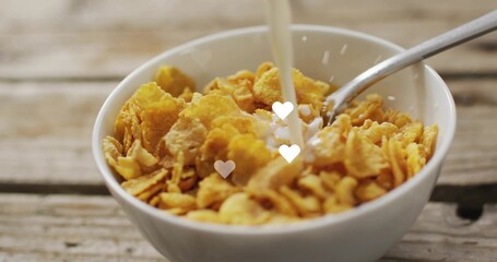 Naklejka premium White ceramic bowl of corn flakes receiving milk pouring on rustic tabletop, spoon and heart bokeh
