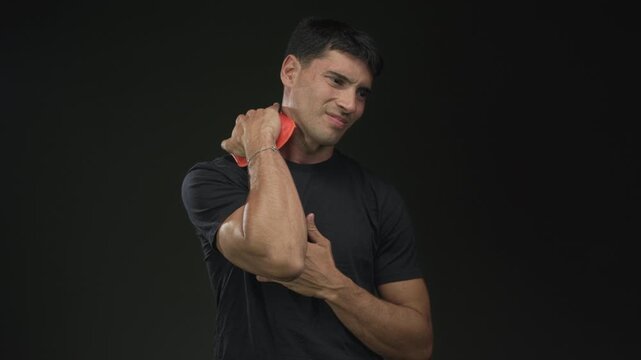 Young hispanic man pressing ice pack to neck in studio with dark green background; pain discomfort healing strain.