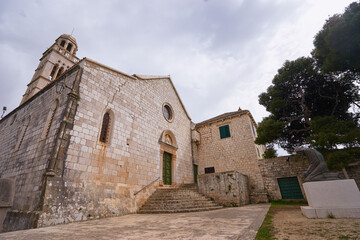 A historic stone church with a bell tower under a cloudy sky, surrounded by trees.