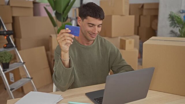 Young man holds creditcard while sitting at laptop inside building amid moving boxes; financial planning confidence.