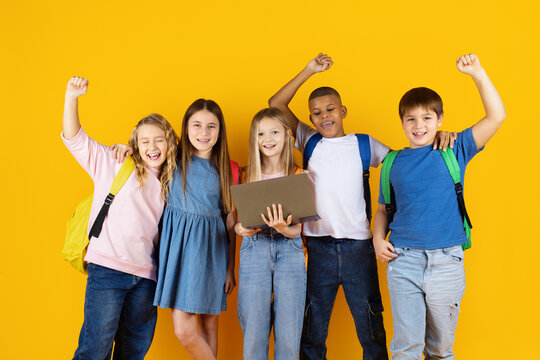 Cheerful group of preteen children with laptop celebrating success with raised arms on yellow studio background. Positive emotions, teamwork, school achievement, and education concept