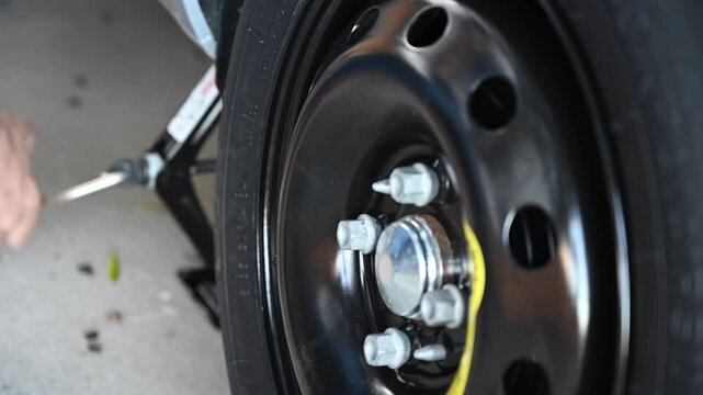 Video of a person using a car jack to lower the vehicle after replacing a flat tire with a temporary spare wheel on the roadside. The scene represents a common roadside emergency situation