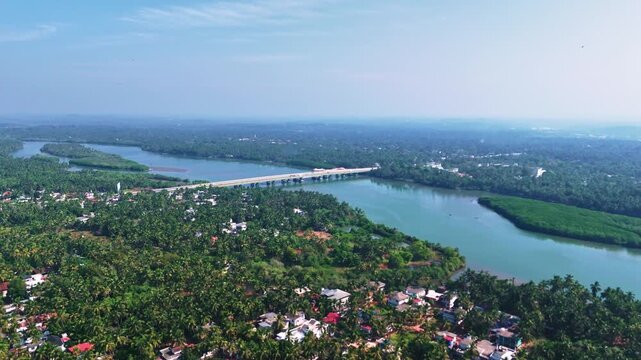 Aerial drone view of Dharmadam showing coastal town layout with lush greenery, houses, roads, and the Arabian Sea in the background.