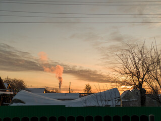 Obraz premium Winter Sunset: Snowy Village with Smoke Plume and Sunset Light