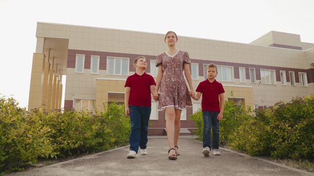 woman walks with two boys in front of a modern building, They hold hands, All three look calm and focused, The boys wear matching red shirts, The woman is dressed in a light summer dress, The building