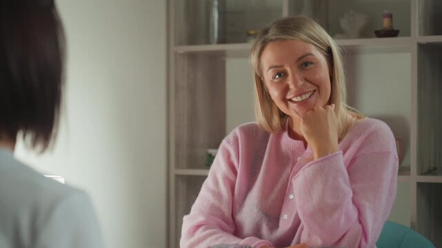 Caucasian woman in pink sweater smiling across table during microcurrent facial consultation with esthetician cozy home spa setting with cups and candle on shelf, soft natural light and warm friendly