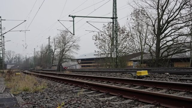 Overcast Rural Railway Tracks With Signal, Empty Rails Stretching Into Distant Horizon Under Gray Sky, Overhead Catenary Wires And Steel Pylons, Leafless Trees And Small Suburban Houses