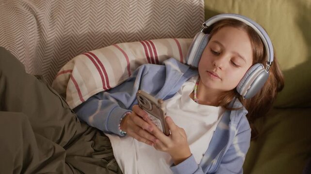 High angle shot of gen Alpha Caucasian girl wearing headphones relaxing on couch at home using smartphone, scrolling newsfeed or texting with friends while listening to music