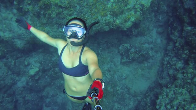 selfie of an asian female woman wearing bikini snorkel mask fin and gloves holding camera shooting from top accenting from underwater facing up to water surface from sea bed coral reef rock day time