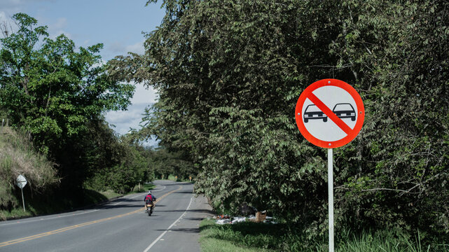 Se&ntilde;al de tr&aacute;nsito de prohibido adelantar en carretera rural con vegetaci&oacute;n y motociclista al fondo
