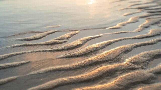 patterns in the sand as water washes over the shore. The sun sets in the distance, creating reflections on the wet surface