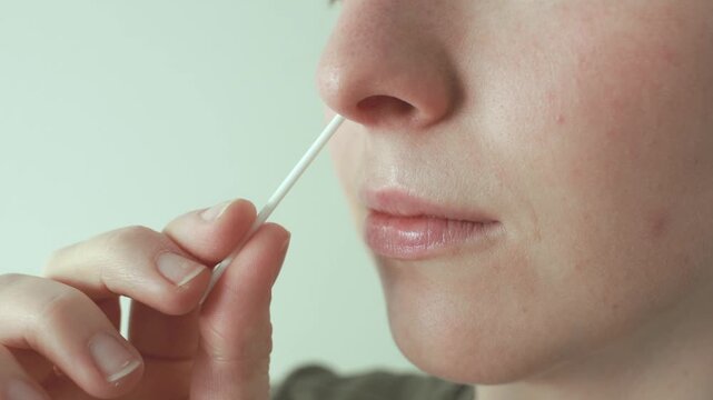 A woman performs a nasal swab for a rapid antigen self-test at home, inserting a swab into her nose to collect a sample. Concept of self testing, healthcare, and disease prevention.