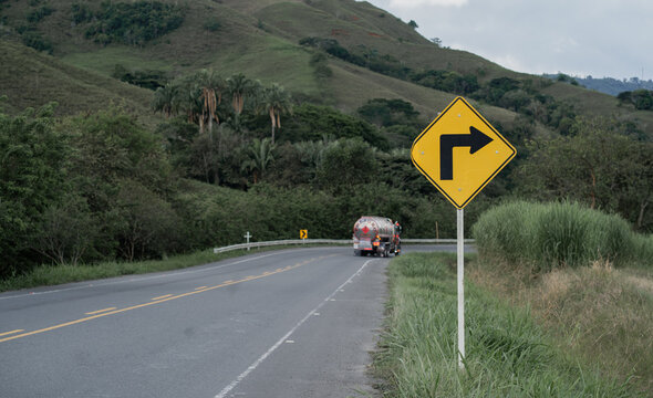 Se&ntilde;al de curva a la derecha en carretera rural con cami&oacute;n cisterna y paisaje monta&ntilde;oso