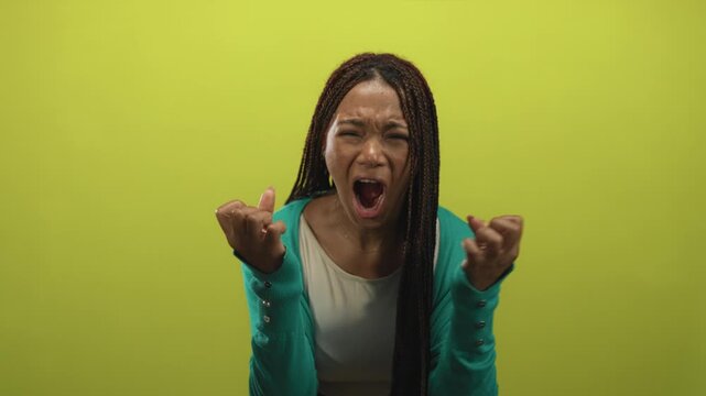 Woman with braided hair shouting, mouth open and hands raised, wearing teal cardigan and necklace in studio with lime green background; anger.