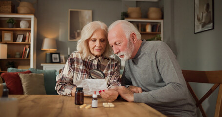 Two adults are sitting at a table in their home. They are reading medication information and...