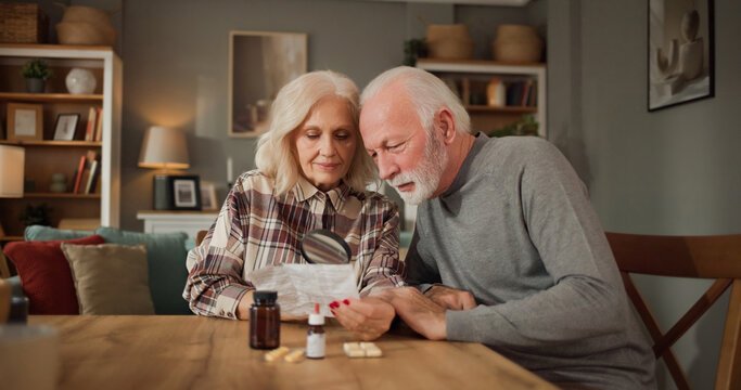 Two adults are sitting at a table in their home. They are reading medication information and discussing the instructions on how to take their medications.
