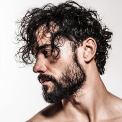 A man stands in a studio with a light background. He looks to the side with curly hair and a full beard