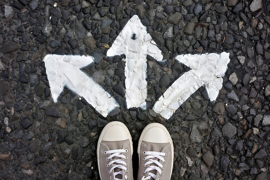 Feet standing on dark asphalt road before white painted arrows pointing in different directions symbolizing important choices and career paths
