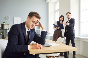 Man at office desk rests head in hand as peers talk by window, with workplace stress, gossip,...