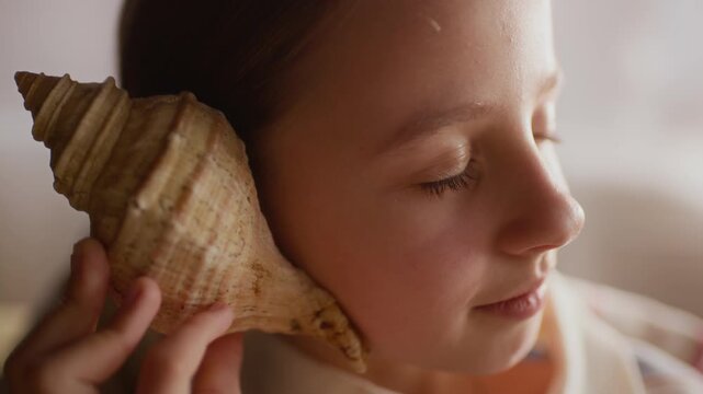 Zoom in shot of dreamy gen Alpha Caucasian girl holding seashell to ear and closing her eyes imagining relaxing sound of ocean
