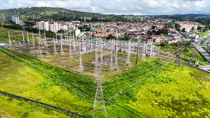 Imagem aérea de subestação e torres de transmissão elétrica com cabos de alta tensão integrados à paisagem urbana e natural. © RNL Fotografia