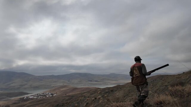 A hunter walking along a river valley on a mountain slope under a cloudy sky. Represents hunting, nature, outdoor lifestyle, rural environment, and human movement within a natural landscape.