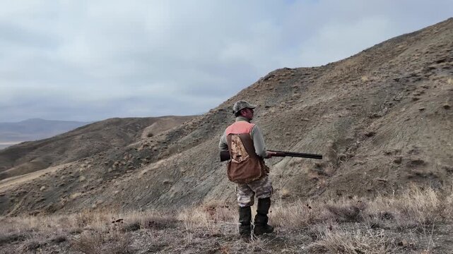Rear view of a hunter walking through dry mountainous terrain carrying a rifle. Represents hunting, nature, outdoor lifestyle, rural environment, and exploration in a documentary-style scene.