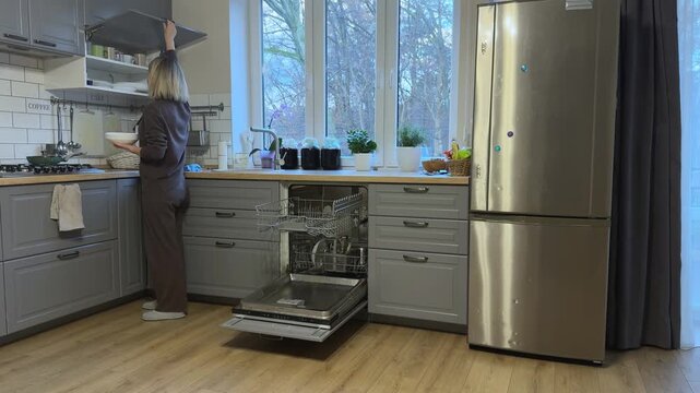 Woman placing clean white plates into upper kitchen cabinet. Daily dish organization and home routine in modern kitchen.