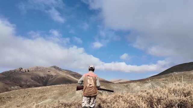 Rear view of a hunter walking through dry mountainous terrain carrying a rifle. Represents hunting, nature, outdoor lifestyle, rural environment, and exploration in a documentary-style scene.