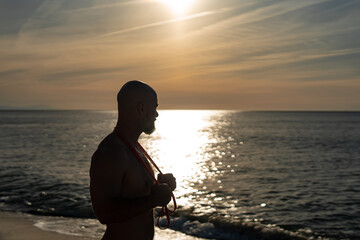 Silhouette of muscular bald man with beard holding resistance band around neck, standing on beach at sunset with golden sunlight reflecting on ocean waves