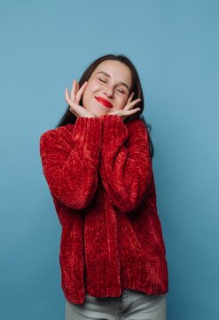 Young woman smiles and poses with hands on her face against a blue background in a casual setting