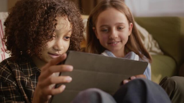 Chest up shot of playful multiethnic gen Alpha girls having fun while using digital tablet, taking turns for watching video or playing game sitting in cozy room by couch