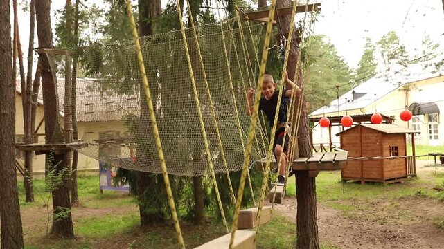 young toddler boy climbing over obstacle with rope in adventure nature activity park. continuous belay system on him. harness and safety line	