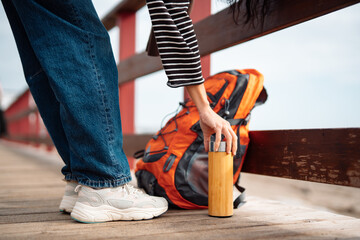 Fototapeta premium Woman hand holding thermos near backpack and sneakers on wooden seaside boardwalk during relaxed outdoor travel lifestyle moment