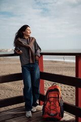 Obraz premium Smiling adult woman standing on wooden beach pier with backpack and smartphone, casual clothing, relaxed posture, coastal background, calm lifestyle moment, outdoor leisure and personal freedom.