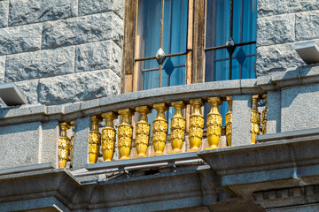 Elegant golden balustrade with ornate columns frames a large window on a rugged stone facade. Sunlight highlights the luxurious details, creating a contrast between opulent decoration and austere arch