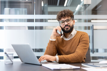 Fototapeta premium Young businessman with curly hair and eyeglasses communicating on his mobile phone while working on a laptop at a contemporary workspace, managing business operations